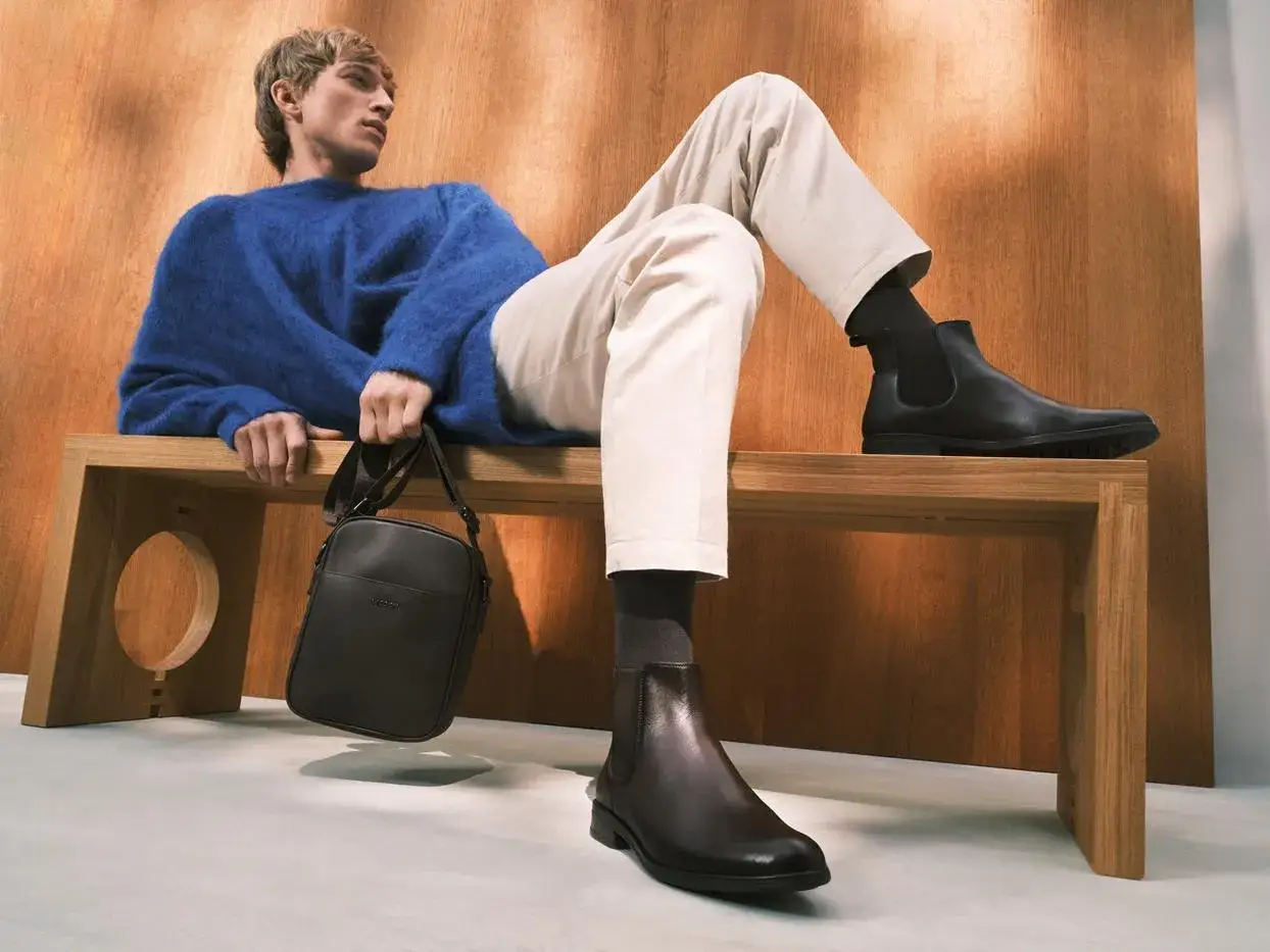 Eleganckie botki to idealne buty do chinosów. Mężczyzna w niebieskim swetrze i beżowych spodniach prezentuje stylową propozycję.