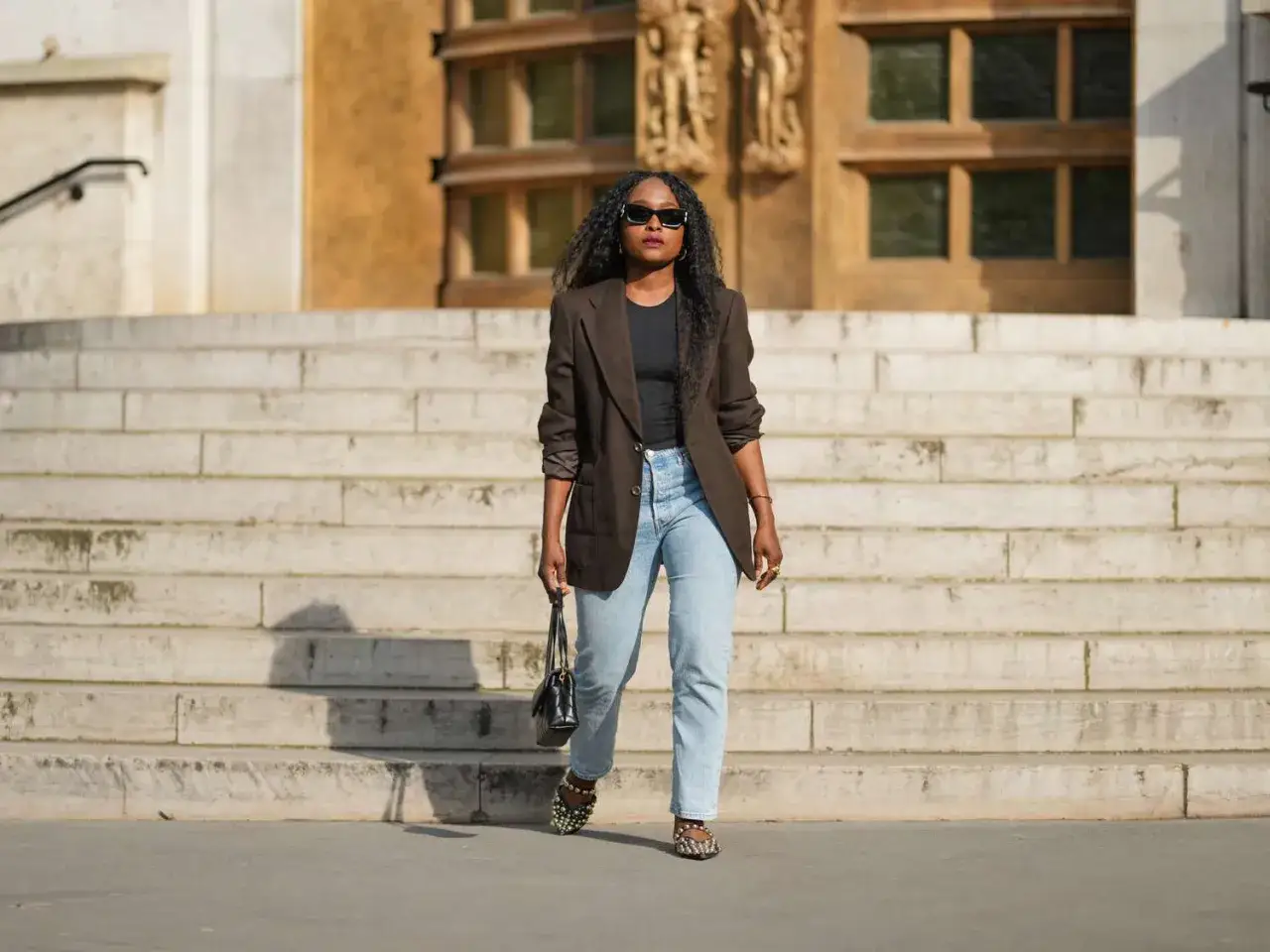 Kobieta w brązowej marynarce i jasnych mom jeans, prezentująca inspirację: jakie buty do mom jeans? Złote sandałki z perełkami.