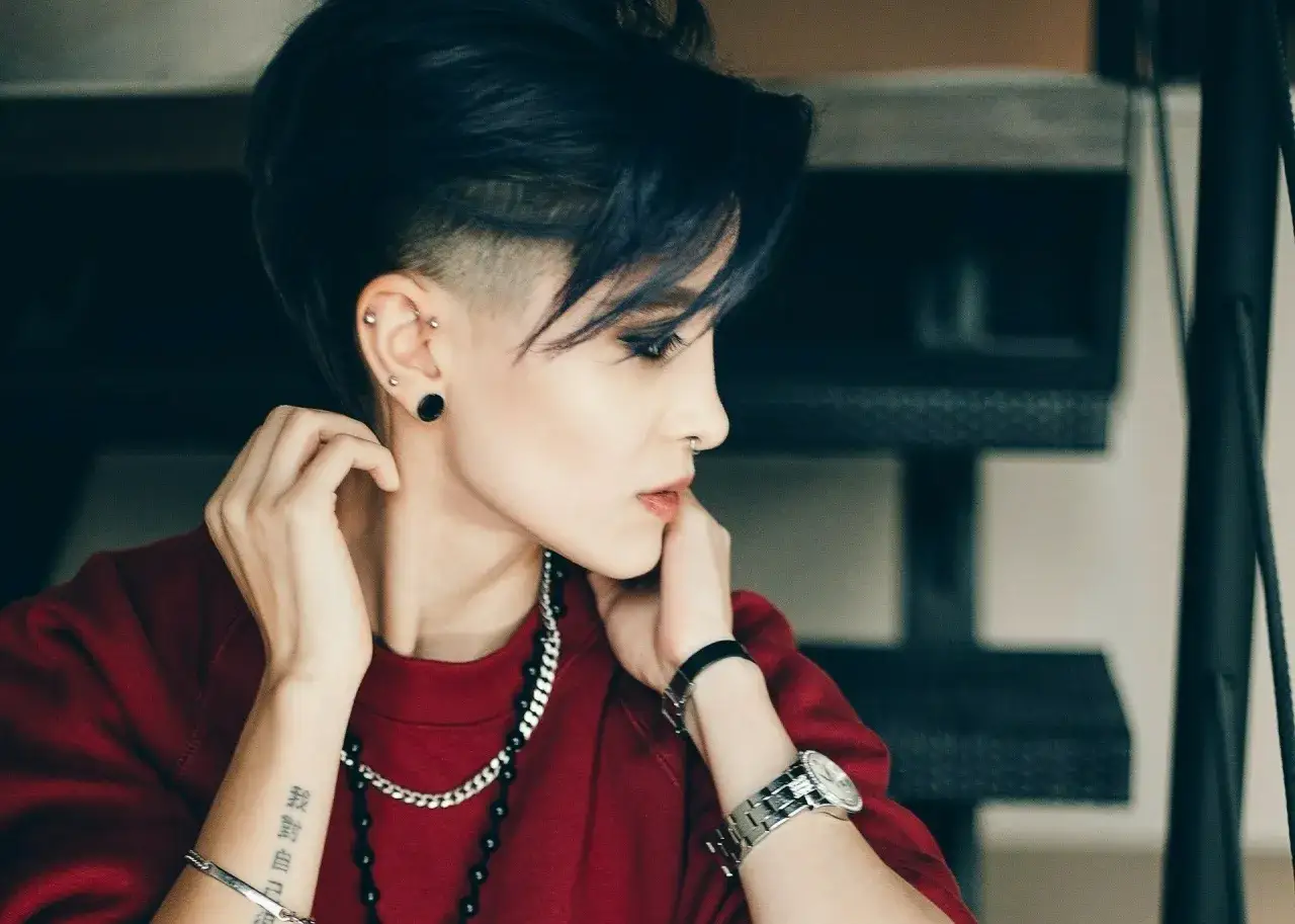 Kobieta z odważną fryzurą buzz cut, w czerwonej bluzie, z licznymi kolczykami i tatuażem na przedramieniu.