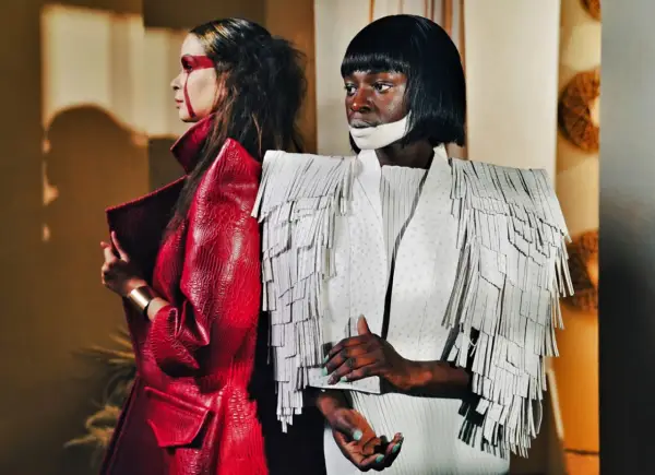 Dwie modelki w awangardowych kreacjach haute couture. Jedna w czerwonej, łuskowej marynarce, druga w białej z frędzlami.